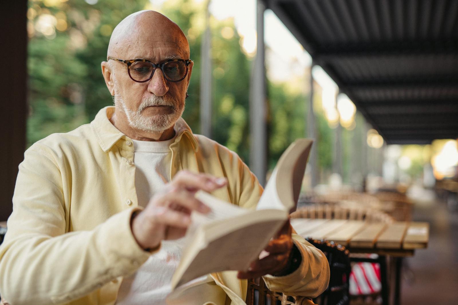 Senior man reading a book outdoors, wearing glasses, in a serene environment.